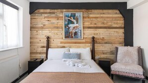 Quarto com parede de madeira, cama com roupa de cama branca, uma cadeira e um quadro.