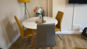 Et rundt spisebord med to stole, en vase med blomster, en lampe og et fjernsyn på væggen.