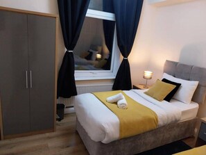 Ein Hotelzimmer mit Bett, Schrank, einem Fenster mit Vorhängen, einer Nachttischlampe und einem Heizkörper.