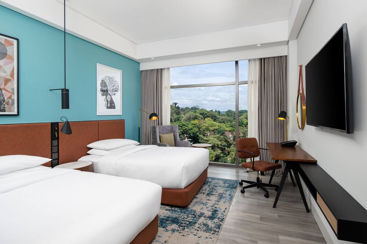 A hotel room with two beds, a desk, a chair, a TV, and a large window with a view of greenery.