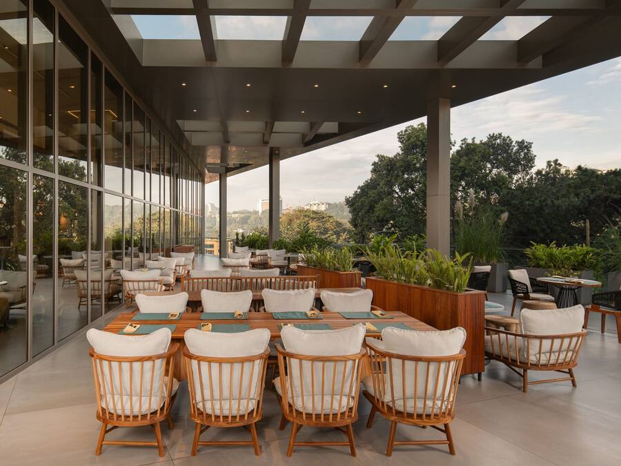 An outdoor dining area with wooden tables and chairs, surrounded by glass walls and potted plants.