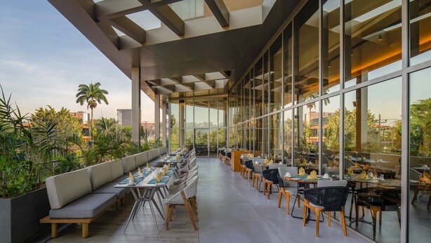 Un área de comedor exterior moderna con un largo banco y mesas, ventanas amplias y vistas de la ciudad.