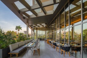 A modern outdoor dining area with a long bench and tables, large windows, and a view of the city. - Four Points by Sheraton Kampala (Kampala)