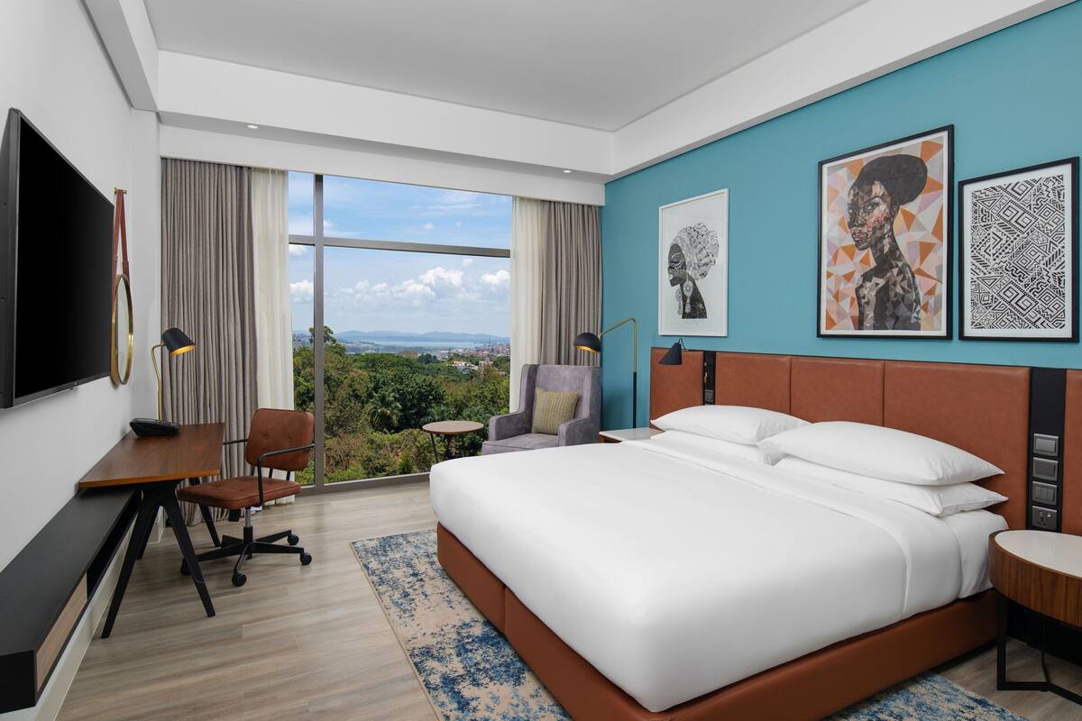A modern hotel room with a large bed, a desk, a chair, and a TV. There are two framed artworks on the wall and a view of the outdoors through large windows.