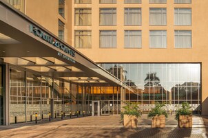 Entrada de hotel con fachada de vidrio, paseo techado y plantas en macetas.