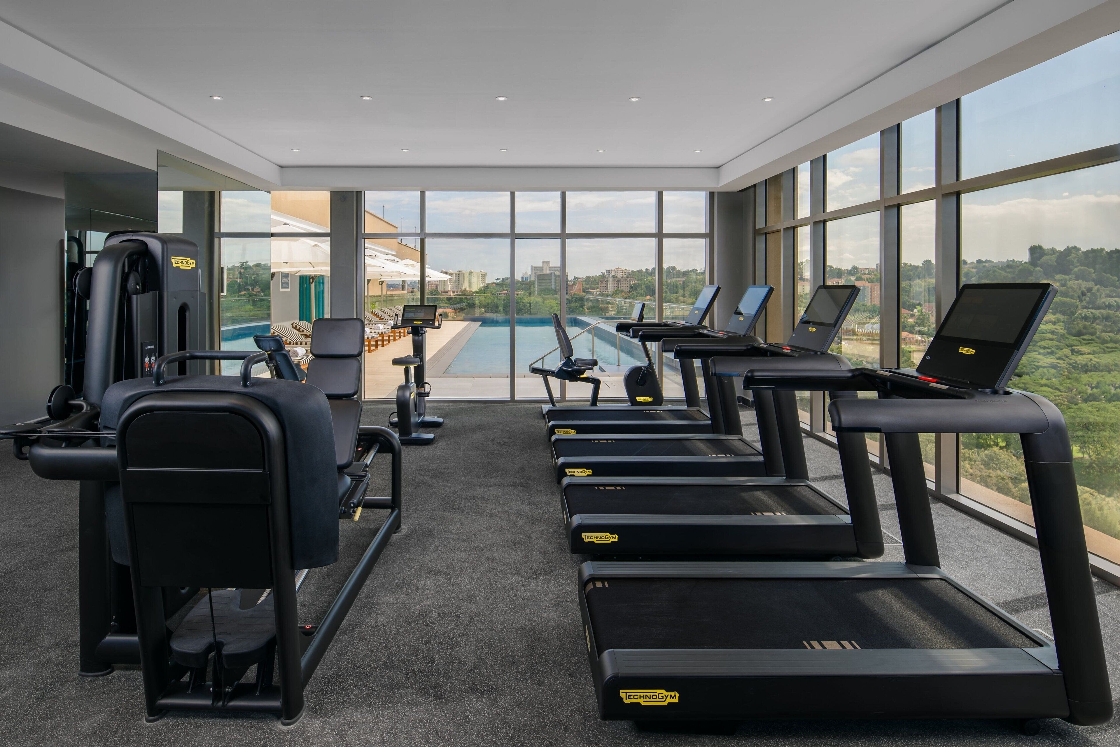 Un gimnasio moderno con cintas de correr, máquinas de pesas y vistas a una piscina y al horizonte urbano.