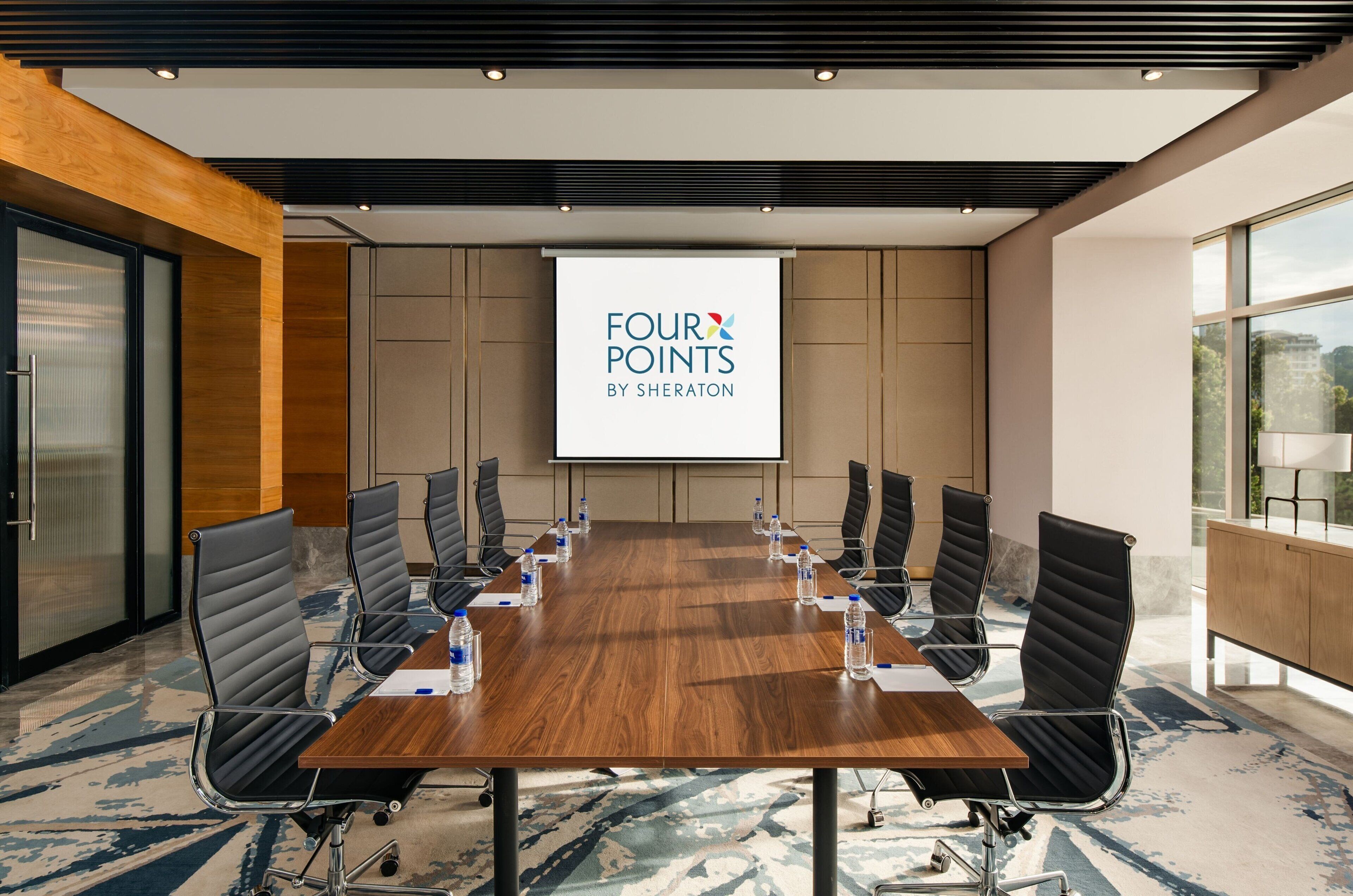 A conference room with a wooden table, black chairs, and a projector screen displaying "FOUR POINTS BY SHERATON".