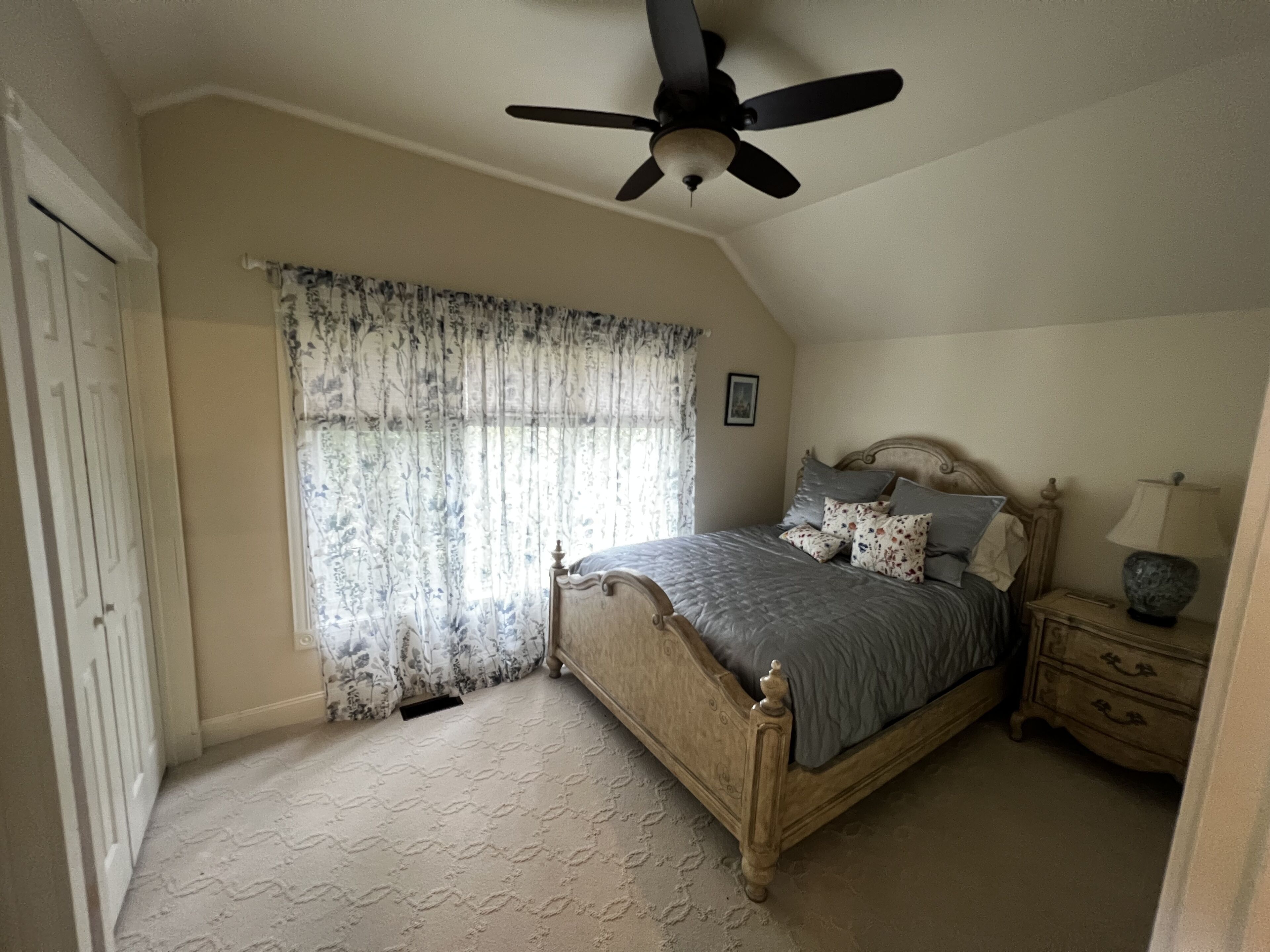 A bedroom with a bed, a nightstand, a lamp, a ceiling fan, a window with curtains, and a door.