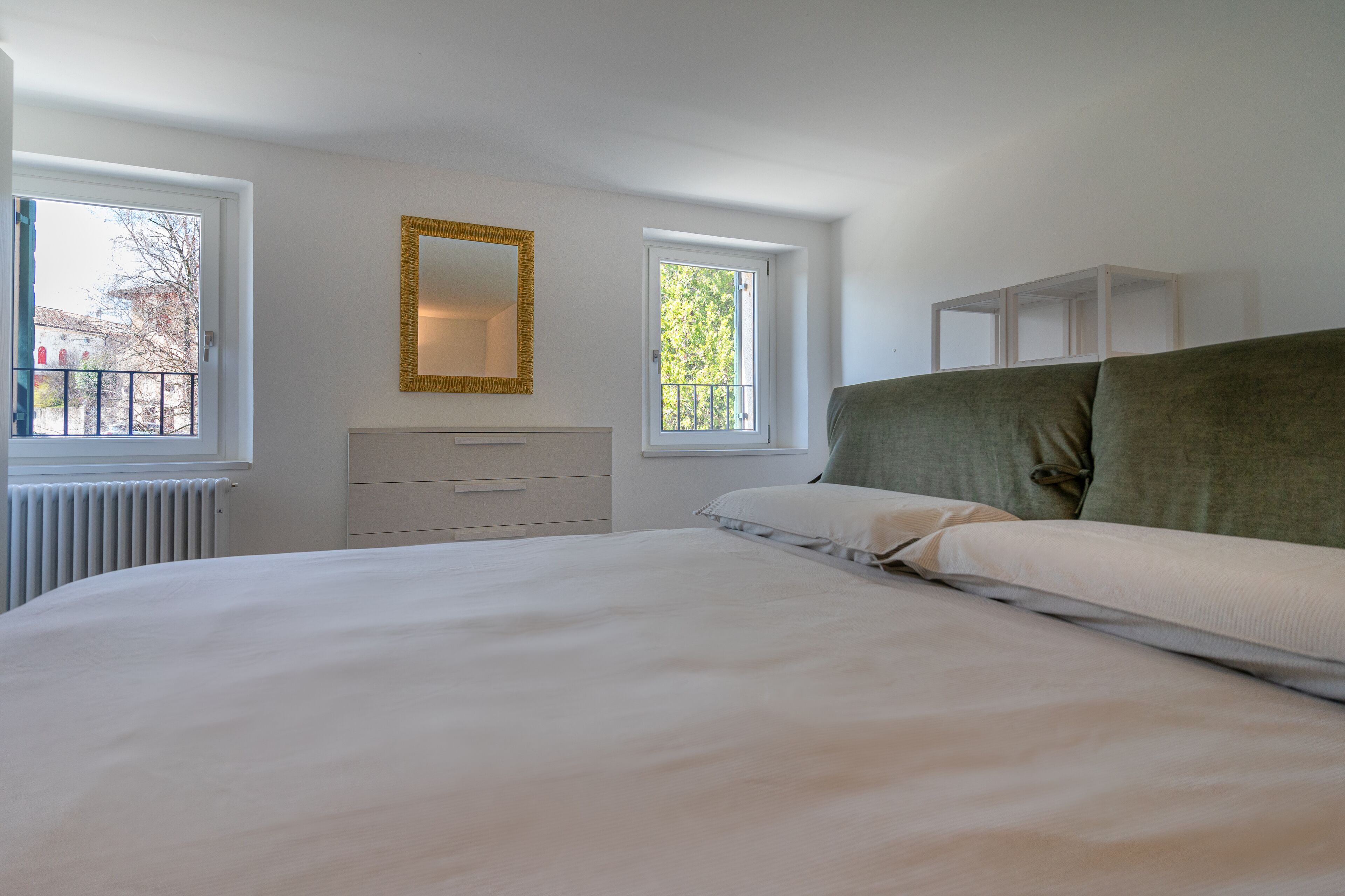 A bedroom with a large bed, two windows, a radiator, a dresser, and a mirror.