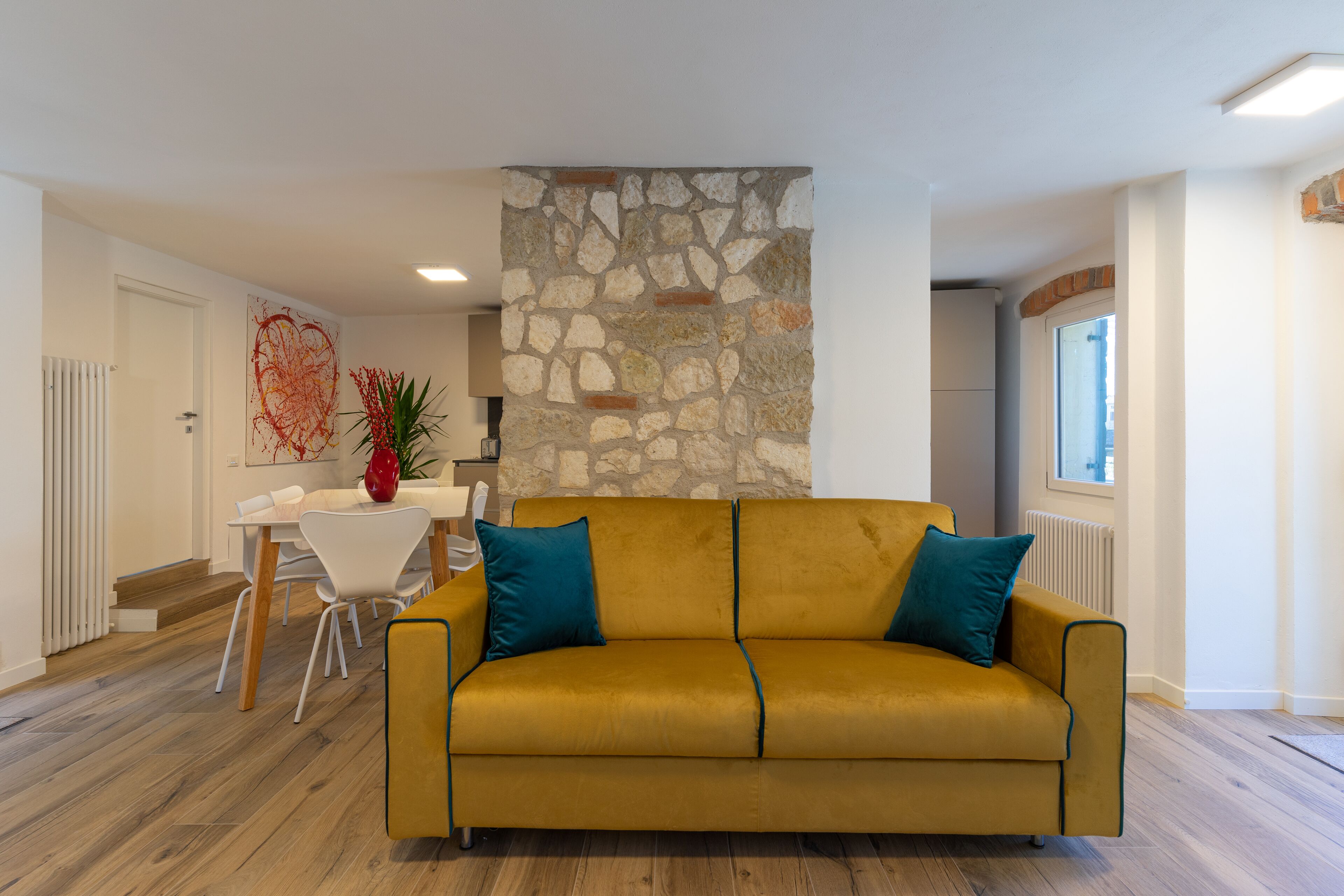 Un salon avec un canapé jaune, un mur en pierre, un coin repas avec une table et des chaises blanches, et une fenêtre sur la droite.