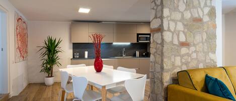 A modern kitchen with a stone accent wall, white dining table with chairs, and a yellow sofa with teal cushions.
