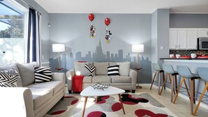 Un moderno salón con un sofá, una mesa de centro y taburetes de bar. Hay un mural con un paisaje urbano en la pared y decoraciones de Mickey Mouse.
