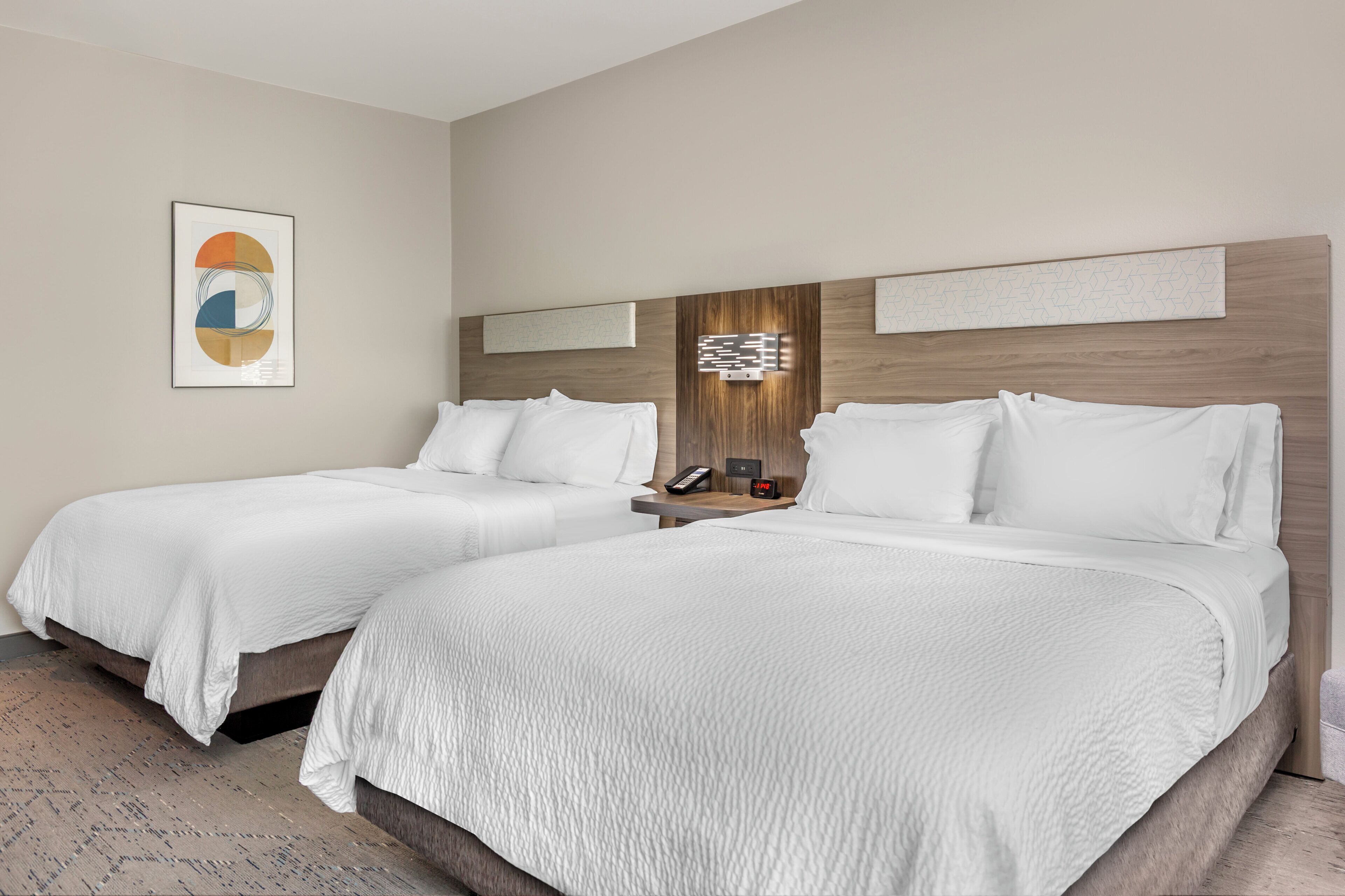 A hotel room with two beds, a nightstand, a wall-mounted phone, and a digital clock.