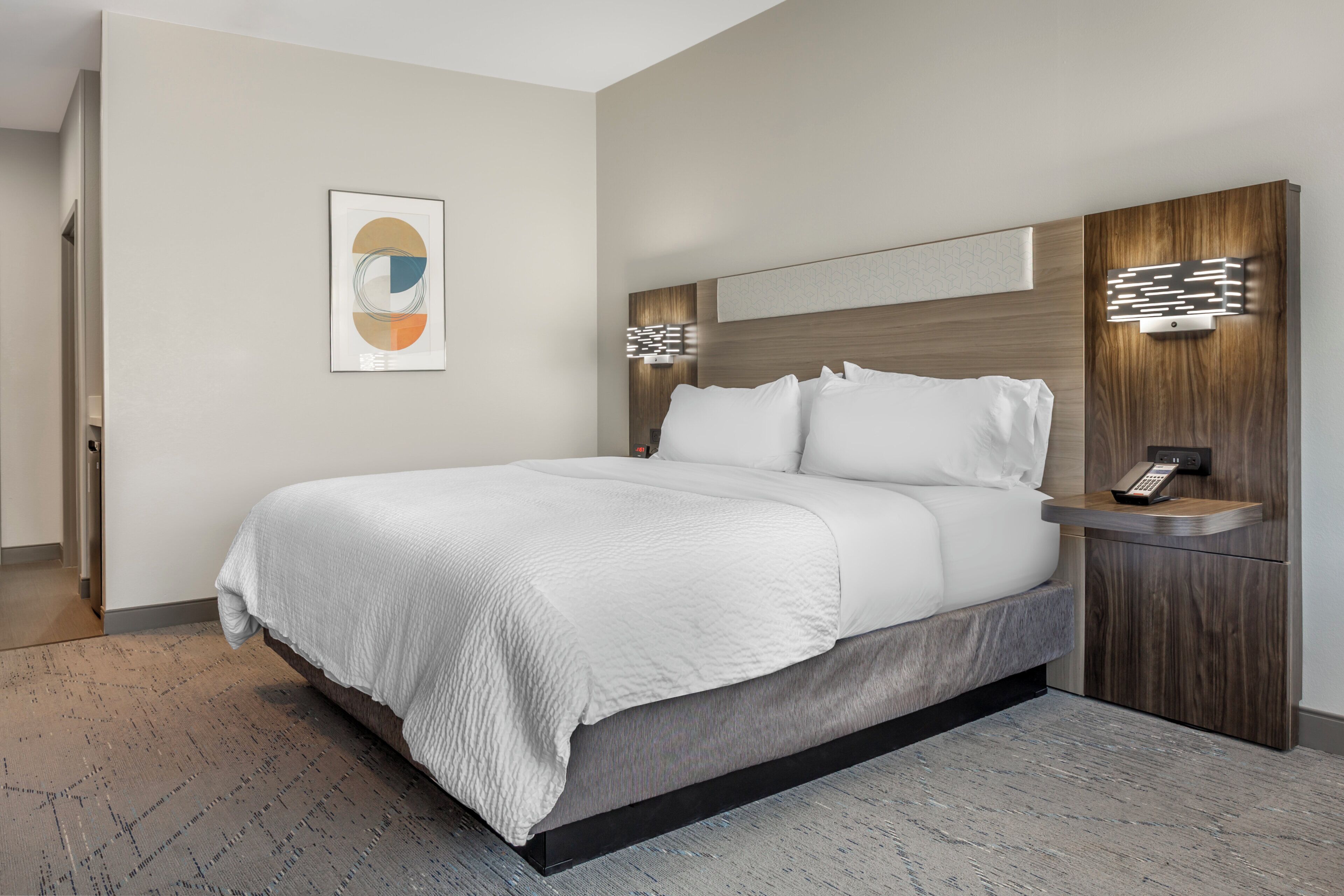 A modern hotel room with a large bed, a bedside table with a phone, and a wall-mounted light fixture.
