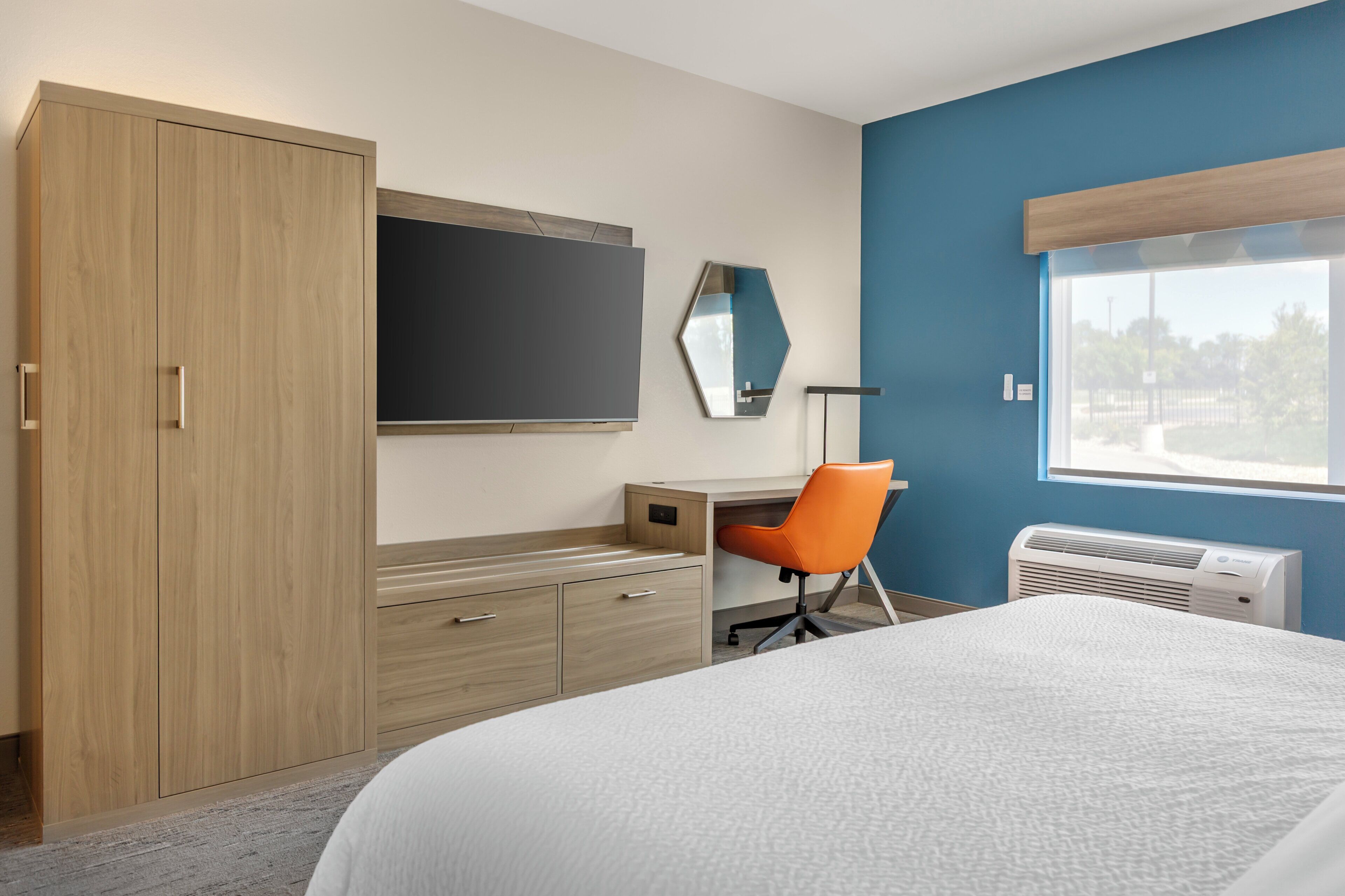 A bedroom with a bed, a desk, an orange chair, a wardrobe, a mirror, and a window with a view.