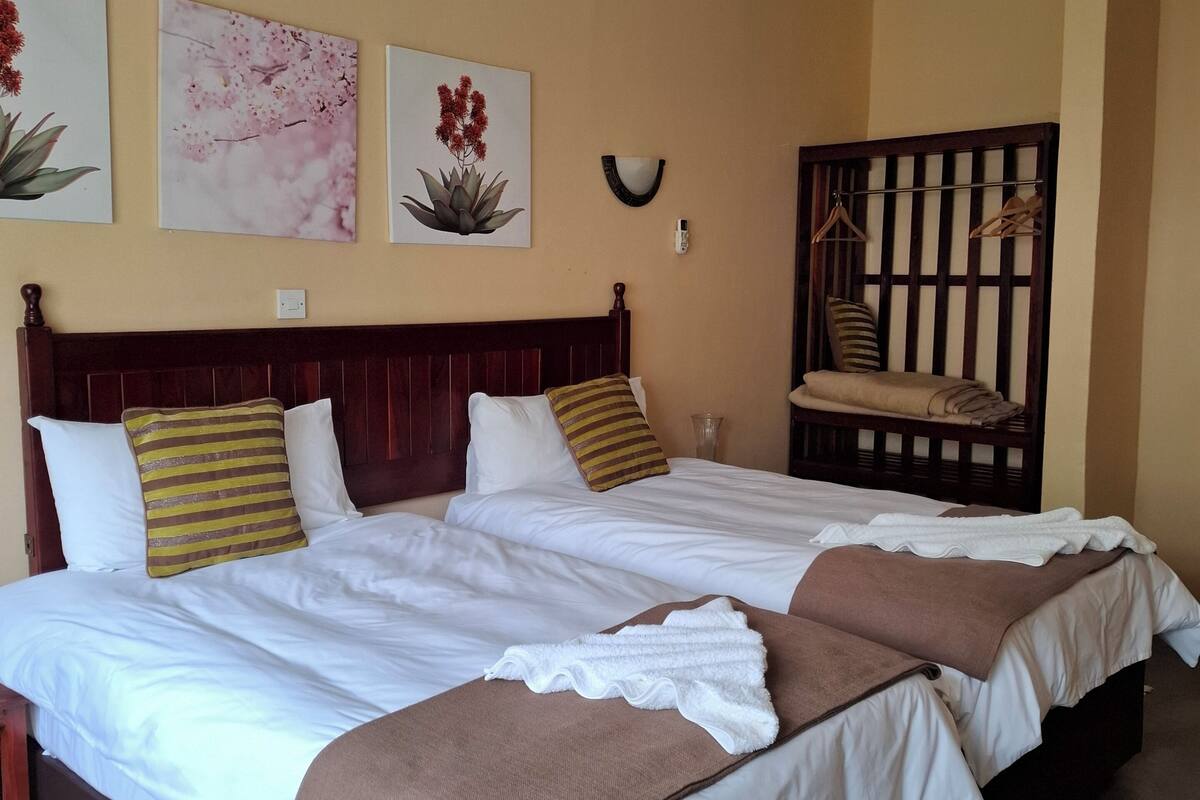 Un lit double avec du linge de lit blanc, deux oreillers et un plaid marron. Trois œuvres d’art représentant des fleurs sont accrochées au mur. Une chaise en bois avec un coussin est placée contre le mur.
