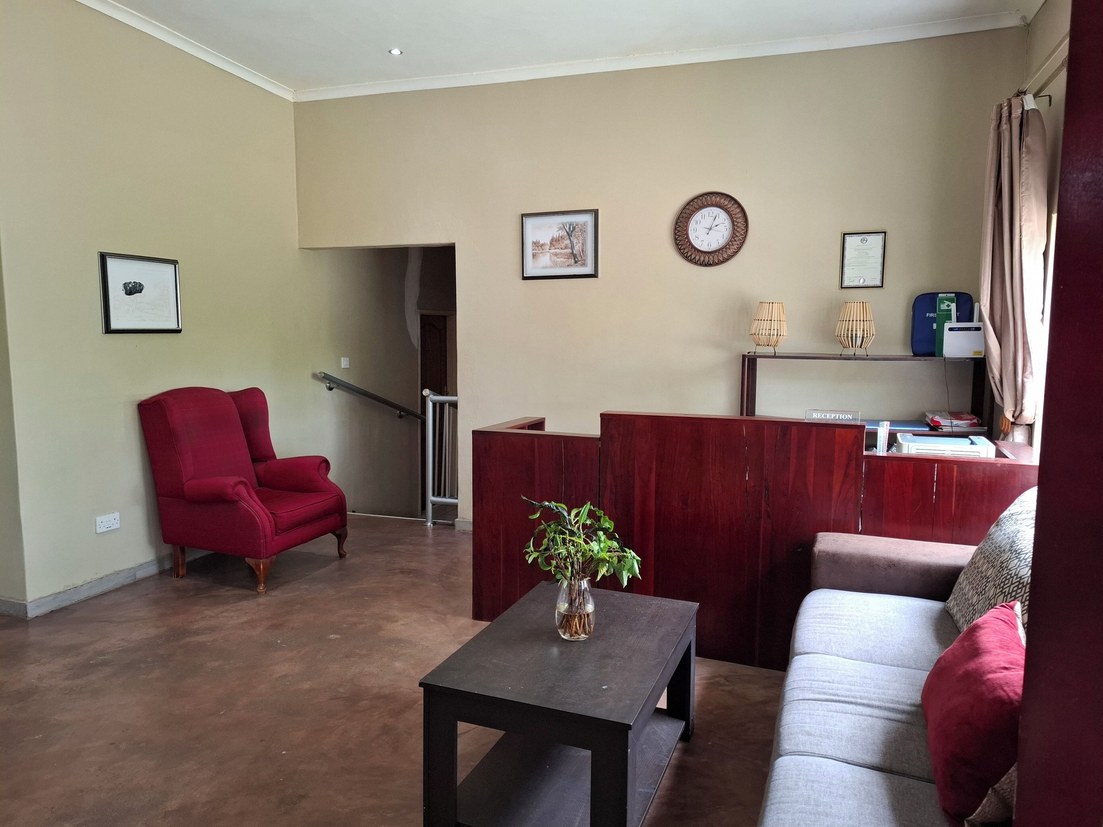 A living room with a red armchair, a grey sofa, a wooden reception desk, and a staircase leading to another room.