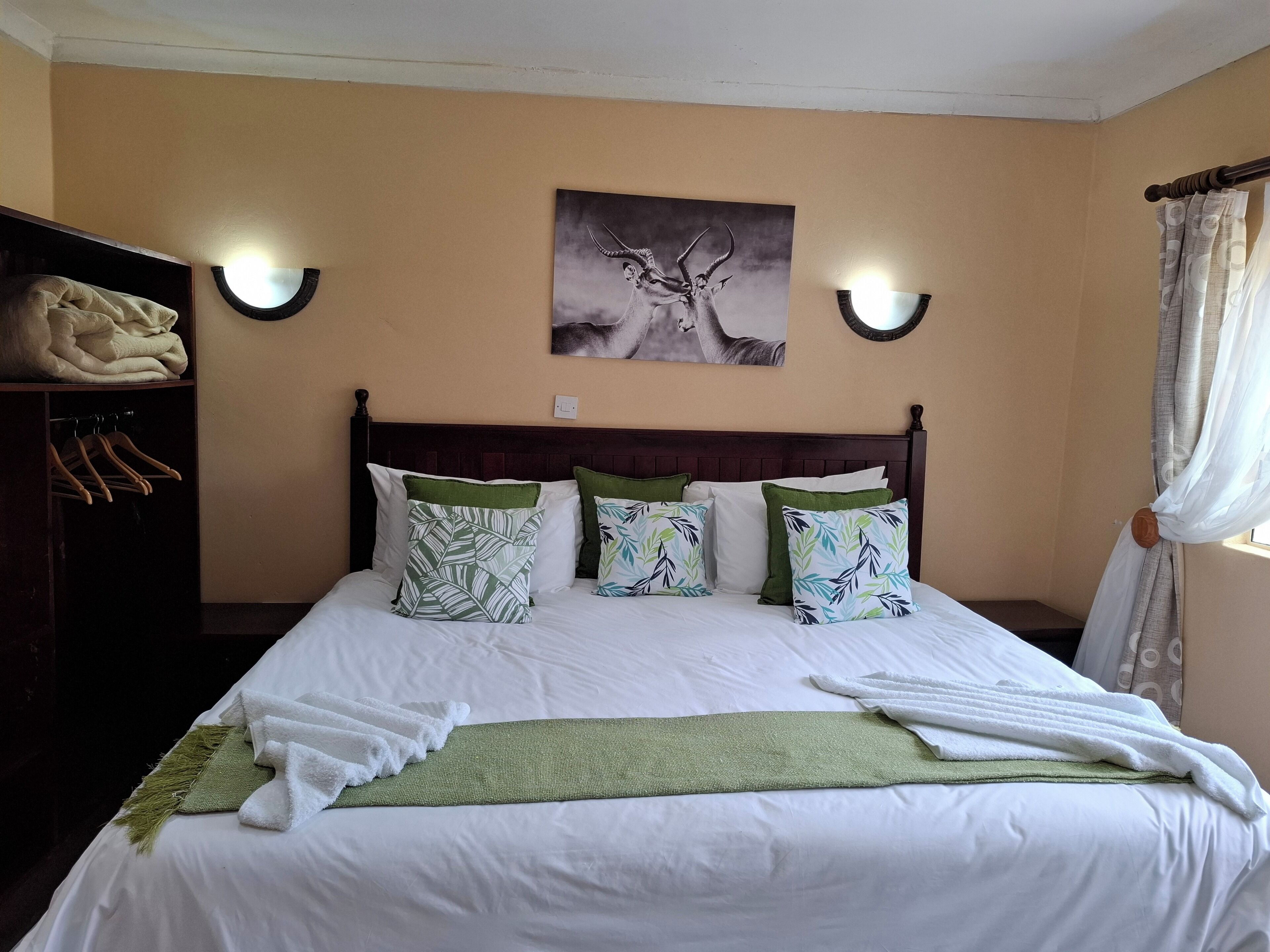 A neatly made bed with white linens and green pillows, a dark wooden headboard, a wall-mounted painting of a deer, and a wardrobe with hangers.