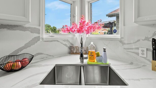 Una cocina con un lavamanos doble, un florero con flores rosadas, un frutero y vistas a una casa a través de las ventanas.