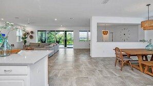 A spacious living area with a large window, a dining table, and a kitchen area with a clock on the wall.