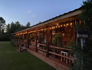 A wooden cabin with a covered patio area, featuring outdoor seating and string lights. - Log Cabin house on 10.5 acers of wooded land. Accommodates 20 plus guest. (Remus)