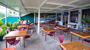 Café al aire libre con mesas y sillas, un mural de una cascada y vista a un puente.