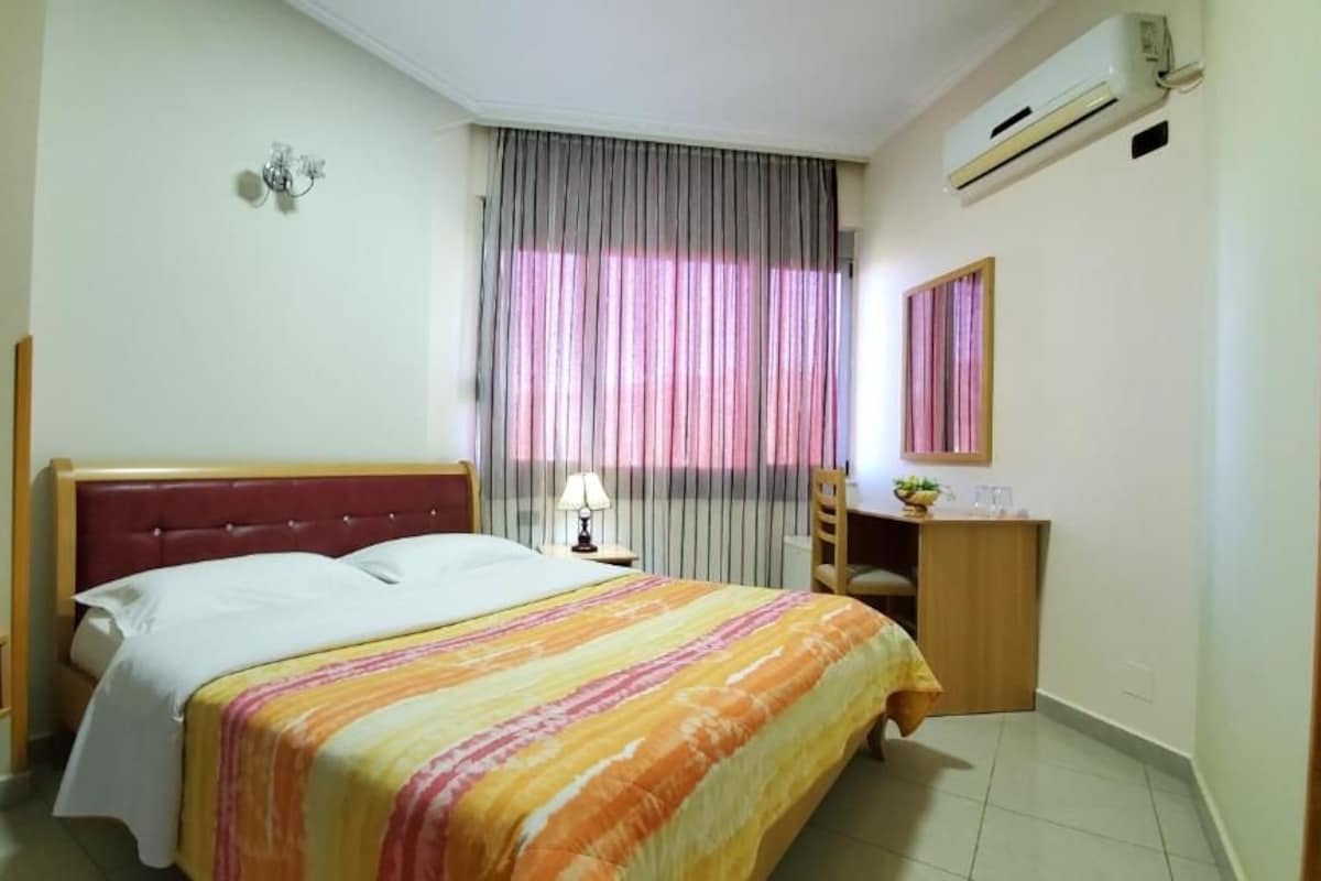 A bedroom with a bed, a nightstand, a lamp, a mirror, and a window with curtains.
