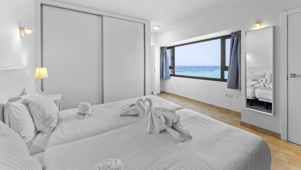 Ein Hotelzimmer mit zwei Betten, einem großen Fenster mit Meerblick, einem Spiegel und einer Wandleuchte.