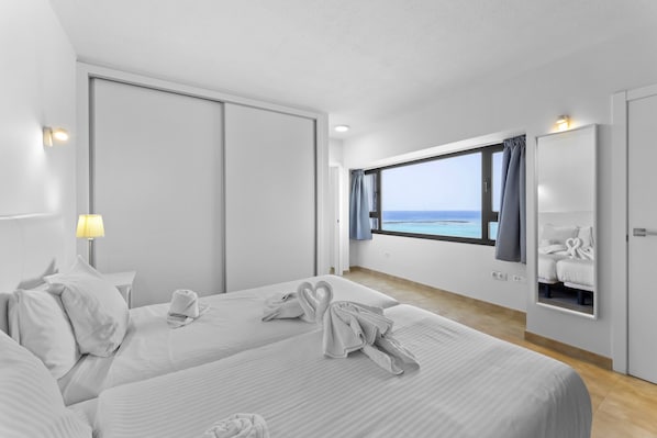 Habitación de hotel con dos camas, un ventanal con vistas al mar, un espejo y una lámpara en la pared.