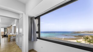 Ein Zimmer mit großem Fenster mit Blick auf einen Strand, Holzboden und einer Tür zu einem weiteren Raum.
