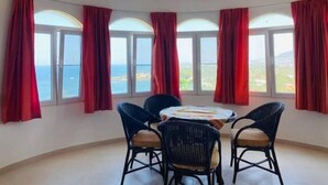 A room with a round table and four chairs, large windows with red curtains, and a view of the sea. - Heavenly View (Cofresi)