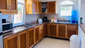 A kitchen with wooden cabinets, a gas stove, a microwave, a sink, and a window with a view of greenery. - Heavenly View (Cofresi)