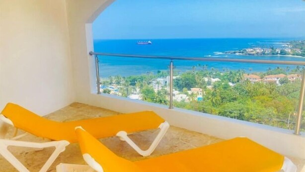 A balcony with two yellow lounge chairs and a view of the sea and buildings. - Heavenly View (Cofresi)