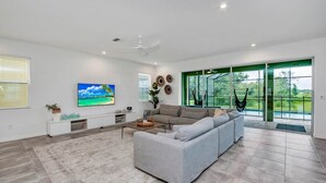A spacious living room with a large sectional sofa, a flat-screen TV, and a large window with a view of greenery. - Solara Savings 2025 9BR Home w/private pool & games for 18 - Clubhouse Included (Four Corners)