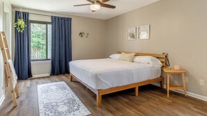 Un dormitorio con cama de madera, una mesita de noche pequeña, un ventilador de techo y una ventana con cortinas.