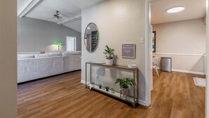 Una sala de estar moderna con piso de madera, una mesa consola de vidrio con plantas, un espejo redondo y un ventilador de techo.