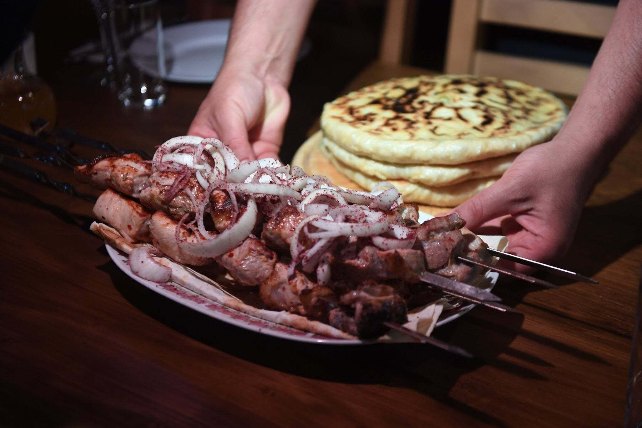 Ein Teller gegrilltes Fleisch mit Zwiebeln und Spießen, dazu ein Stapel Fladenbrotes auf einem Holztisch.