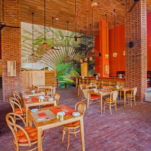 Un restaurant avec des tables et des chaises en bois, une fresque représentant des feuilles tropicales et des lampes suspendues.