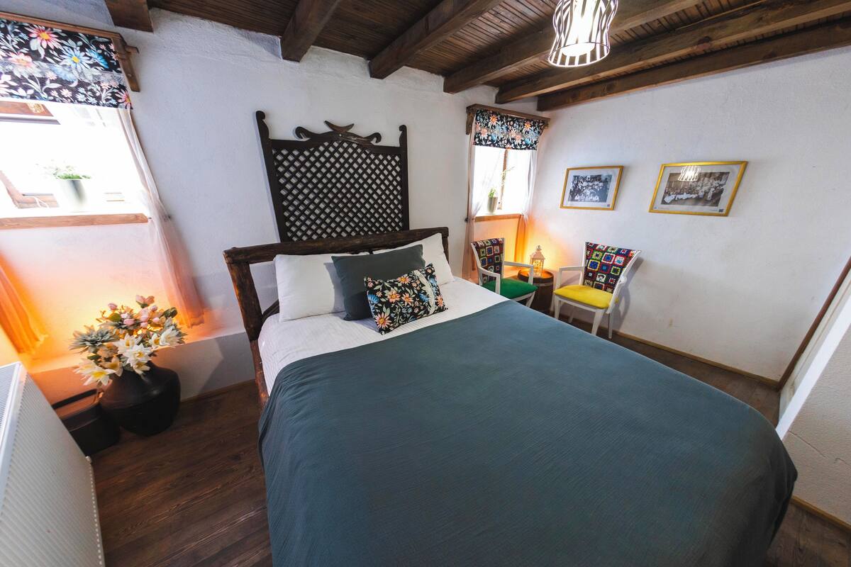 A bedroom with a bed, a wooden headboard, a chair, a small table, and a wall with framed pictures.