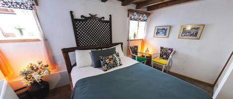 A bedroom with a bed, a wooden headboard, a chair, a small table, and a wall with framed pictures.