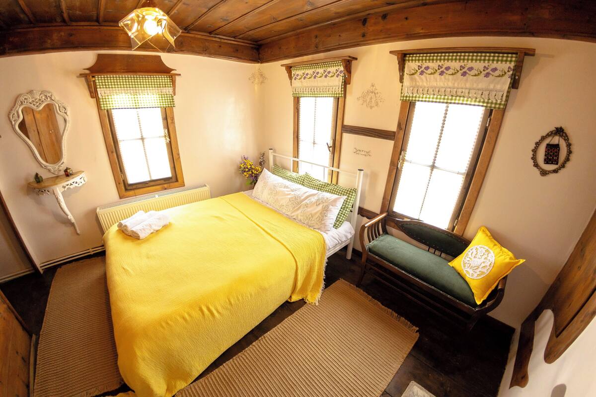 A room with a bed, a chair, a window with checkered curtains, and a wall-mounted mirror.