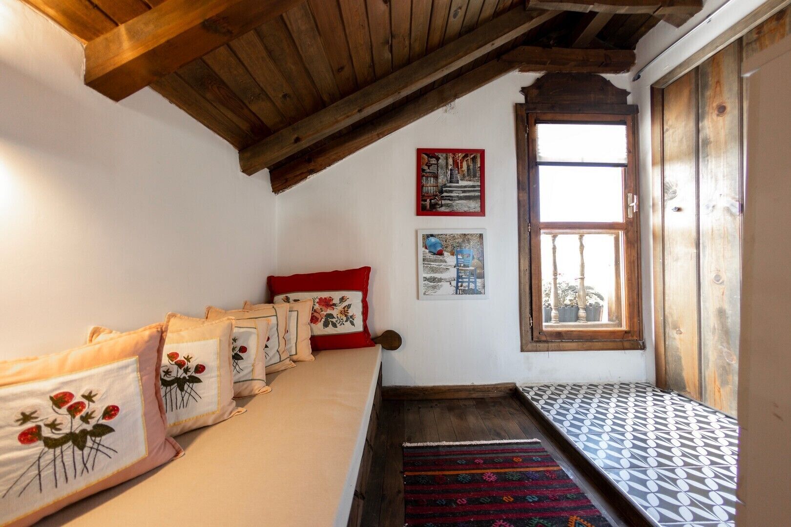 A cozy room with a wooden ceiling, a sofa with floral pillows, a red cushion, and a patterned rug.