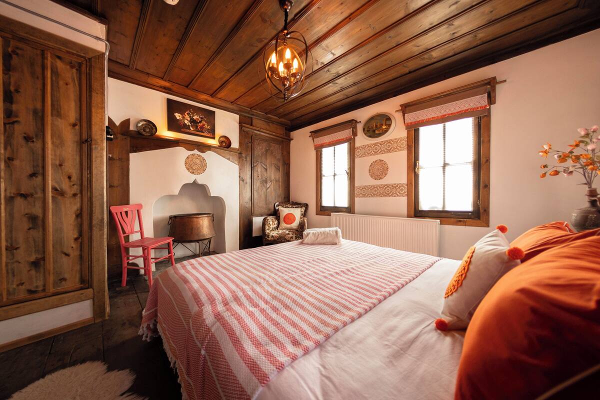 A bedroom with a wooden ceiling, a bed with a striped bedspread, a red chair, and a window with decorative shutters.