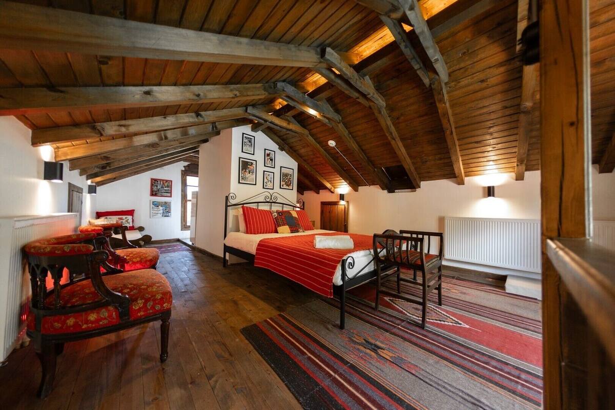 A cozy room with a wooden ceiling, a bed with a red blanket, a wooden chair, and a patterned rug.