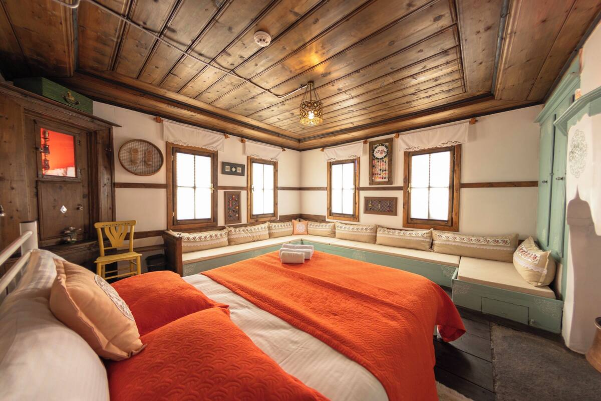 A traditional room with a wooden ceiling, a large bed with an orange bedspread, a bench with cushions, and a green cabinet.