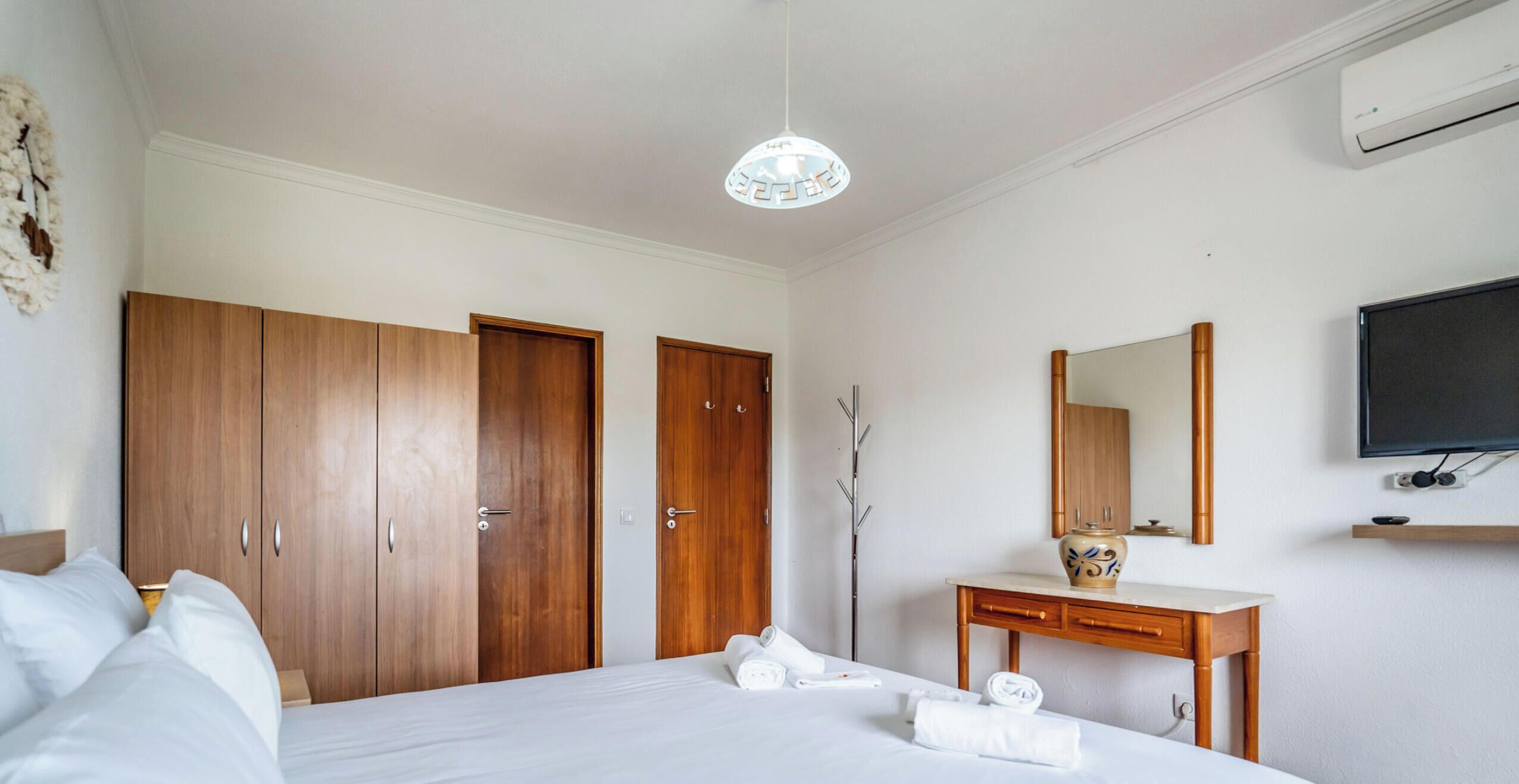 A hotel room with a bed, a wooden wardrobe, a desk with a mirror, and a television.