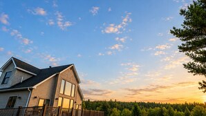 A two-story house with a deck, surrounded by a grassy yard and a forested area.