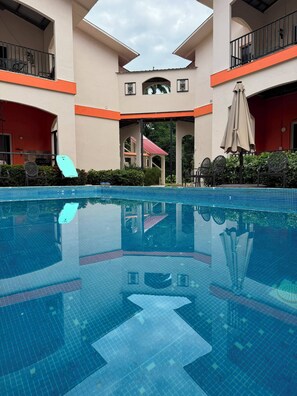 Ein Schwimmbecken mit Fliesenboden, umgeben von einem Gebäude mit Balkonen und einem überdachten Bereich mit Stühlen und Sonnenschirm.