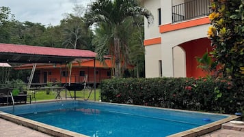 A swimming pool with a tiled deck, surrounded by greenery and a building with a balcony.