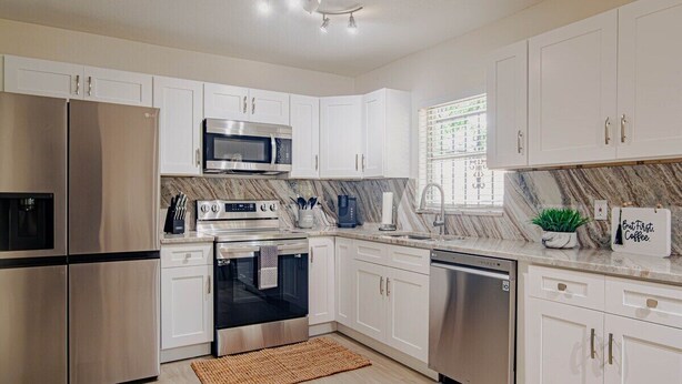 A modern kitchen with white cabinets, stainless steel appliances, a marble backsplash, and a window with blinds.